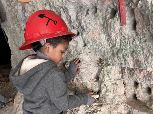 A small child wearing a red hard hat touches a rock wall inside a rocky space.