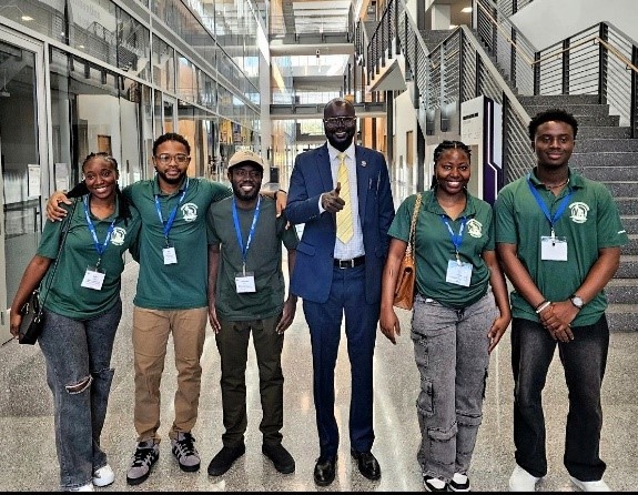 Six people wearing matching green polo shirts and name badges stand with a man in a navy suit and yellow tie inside a modern building with glass walls and staircases, smiling and posing together for a group photo.