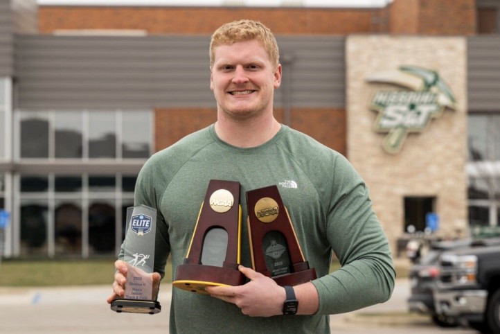 A smiling man stands outdoors in front of a university building, holding three awards, including two wooden trophy plaques and a clear glass trophy, wearing a green long-sleeve shirt with the NCAA Elite 90 logo.
