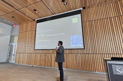 A person stands on a stage in a modern auditorium, holding a microphone and presenting in front of a large projected slide with text and a QR code, surrounded by wood-paneled walls.