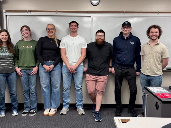 Eight people stand shoulder to shoulder in a classroom in front of a whiteboard, smiling at the camera. They are dressed casually in jeans and sweaters or T-shirts, with a clock above the whiteboard and classroom desks visible in the foreground.