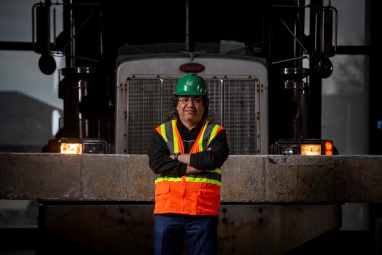 A construction worker wearing a green hard hat and an orange high-visibility safety vest stands with arms crossed in front of a large truck, positioned between its headlights in a dimly lit industrial setting.