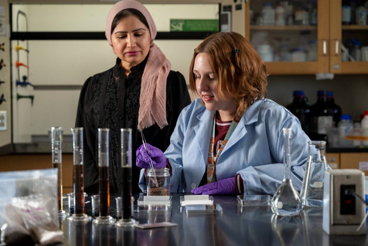 In a science laboratory, two women work at a lab bench with glassware and measuring instruments. One woman in a light blue lab coat and purple gloves carefully pours liquid into a small container, while another woman wearing a pink headscarf and dark clothing observes closely from beside her.