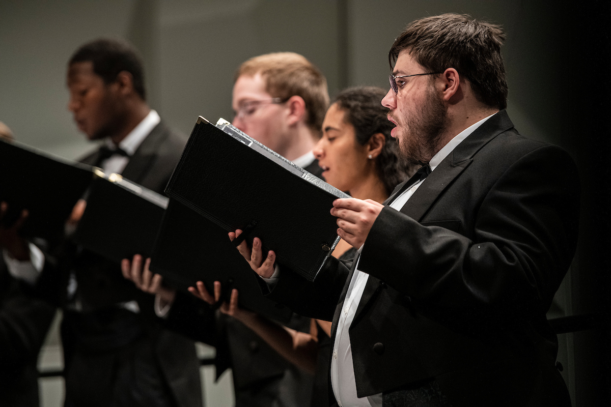 Students wearing suits and ties hold black folders and sing from sheet music.