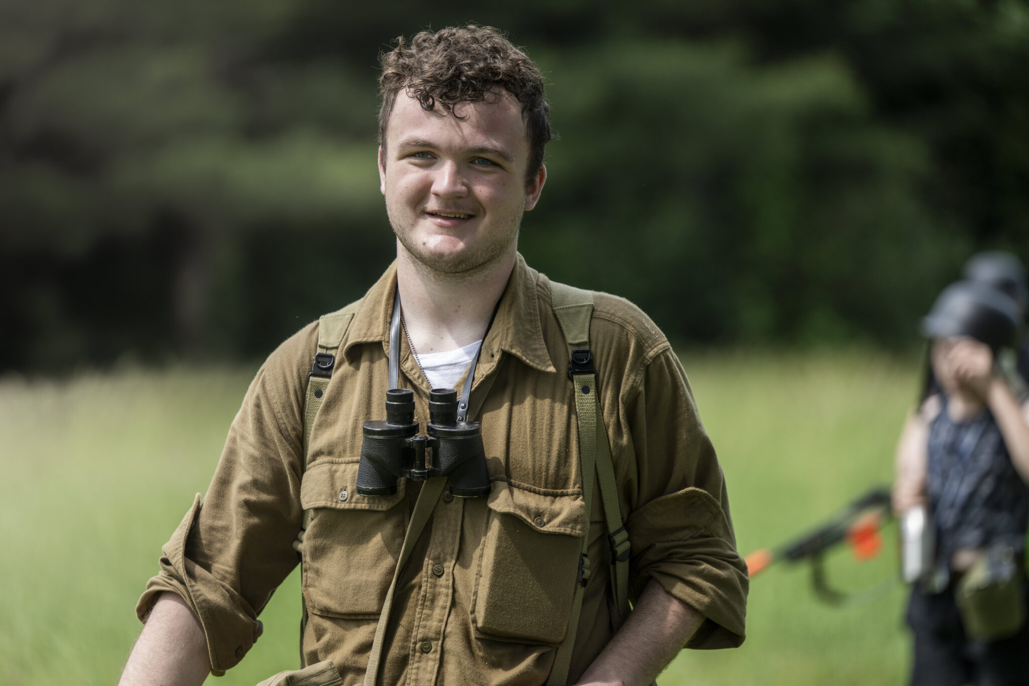 A person wearing a brown shirt and binoculars smiles while standing in a grassy field, with others blurred in the background.