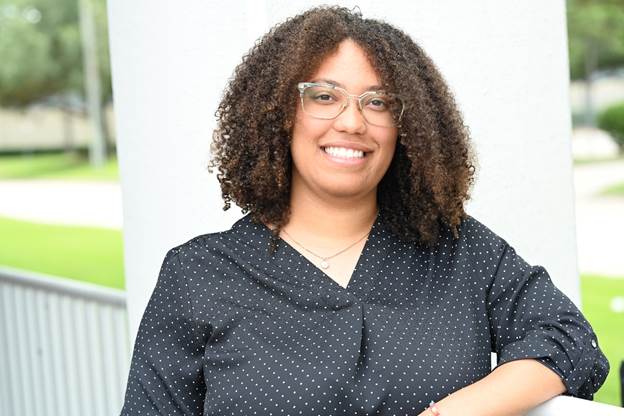 A smiling woman with curly hair and glasses wearing a black polka‑dot blouse, standing outdoors near a railing.