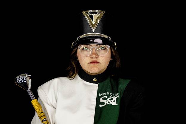 Person wearing a Missouri S&T marching band uniform and hat, holding a ceremonial mace, facing the camera against a black background.