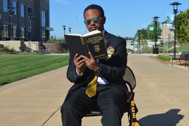A person wearing a black suit and sunglasses sits on a chair along a campus walkway, reading a book on a sunny day.