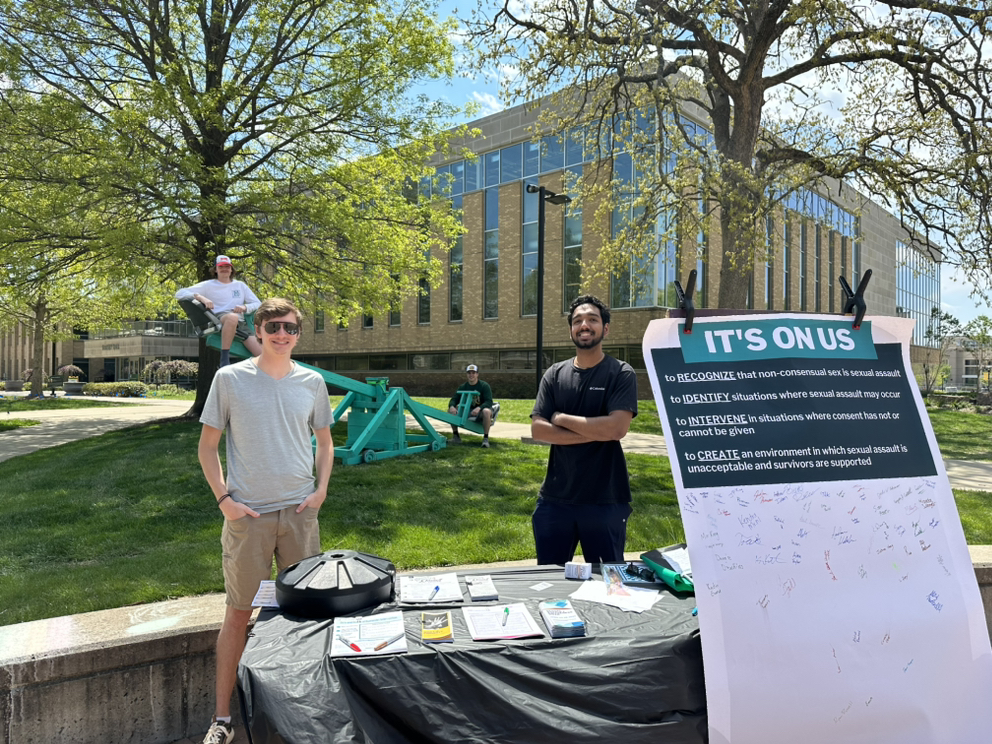 Two people stand behind an information table on a sunny campus lawn, with an “IT’S ON US” sign, flyers, and a poster board; trees, students, and an academic building are in the background.