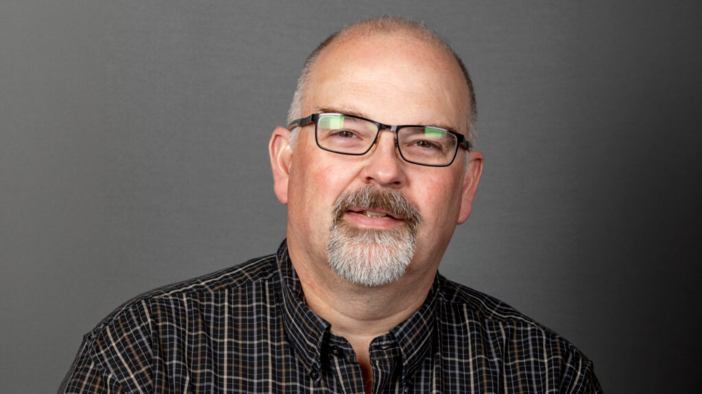 Close-up portrait of a person wearing glasses and a checkered shirt against a gray background.