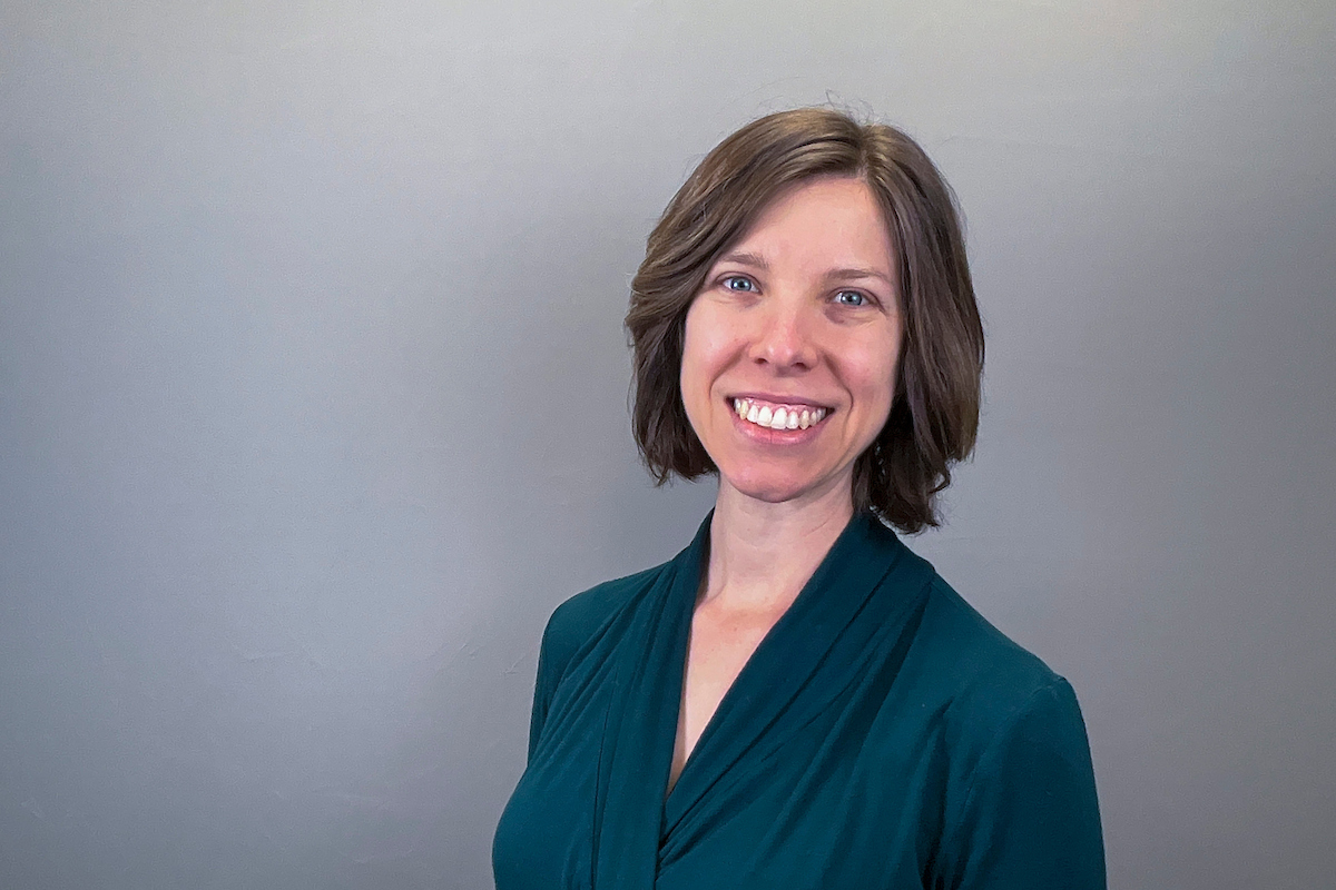 Person with short brown hair wearing a teal blouse, smiling toward the camera against a plain gray background.