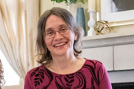 Person with glasses wearing a red patterned top, seated indoors near a fireplace and decorative shelves.