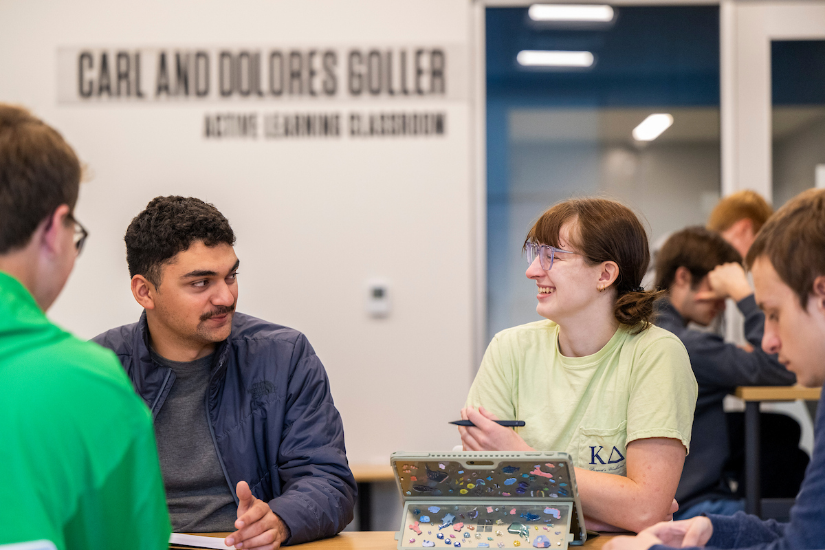 Group of students sitting at a table, talking and working together, with laptops and notebooks; a sign for “Carl and Dolores Coller Active Learning Classroom” is visible in the background.