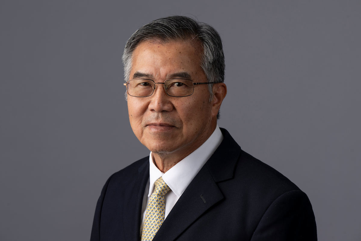Person wearing glasses, a dark suit, and a patterned tie, posed against a plain gray background.