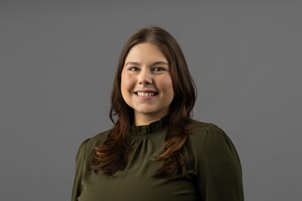 Person with shoulder-length hair wearing a dark green top, posing against a plain gray background.