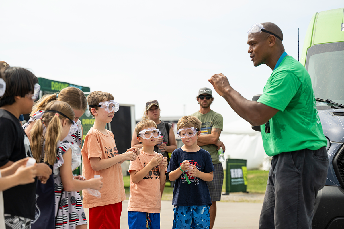 Children wearing safety goggles watch an instructor lead an outdoor science activity.