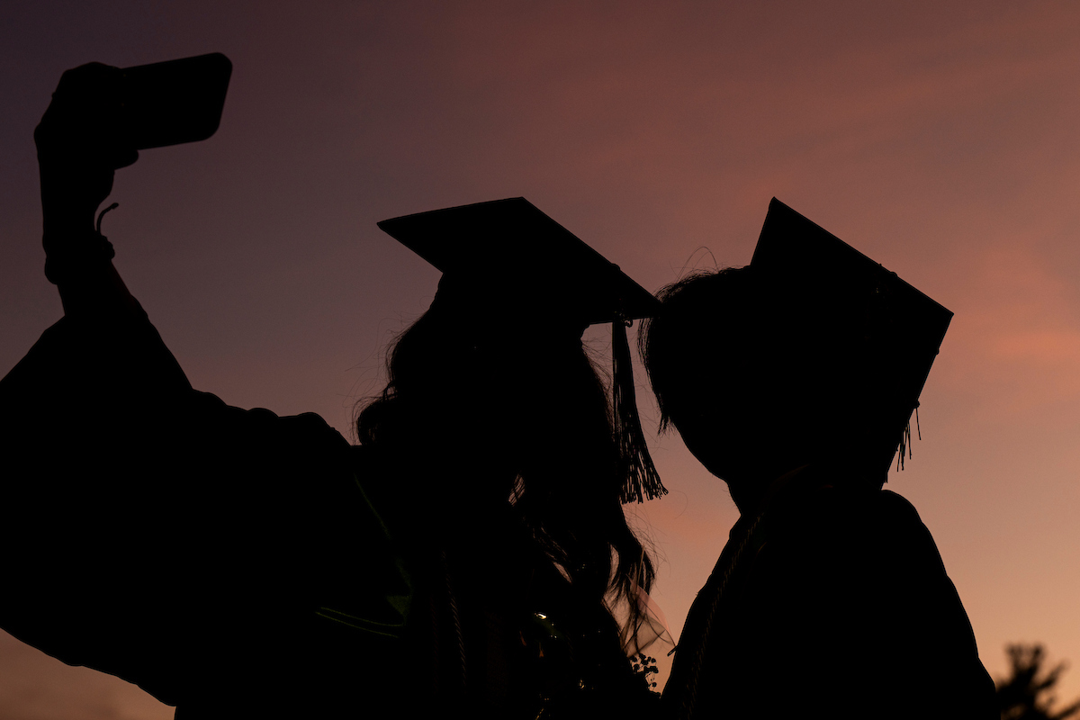 Silhouettes of two graduates in caps taking a selfie against a sunset sky.