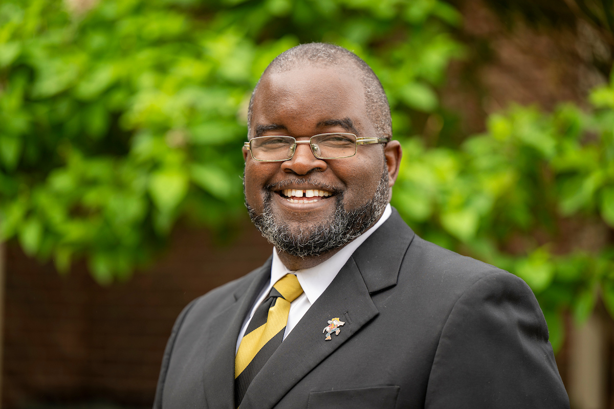 Portrait of a person wearing glasses, a black suit, white shirt, and black-and-yellow striped tie with a small lapel pin, standing outdoors in front of green foliage; the person is facing the camera with a broad smile.