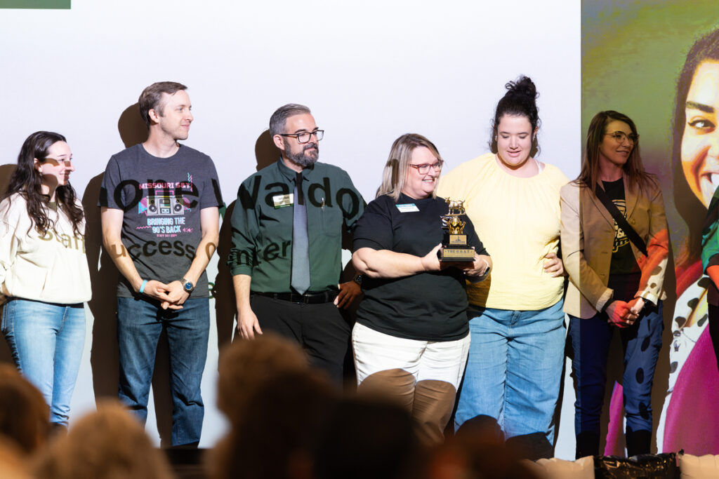 Group standing on stage while one person holds a trophy award.
