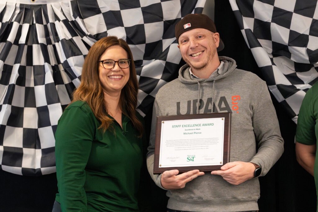 Two people on stage holding a “Staff Excellence Award” certificate at S&T’s Staff Appreciation Day event.