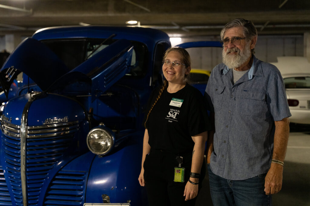 Two people standing beside a vintage blue truck with its hood open in an indoor display area.