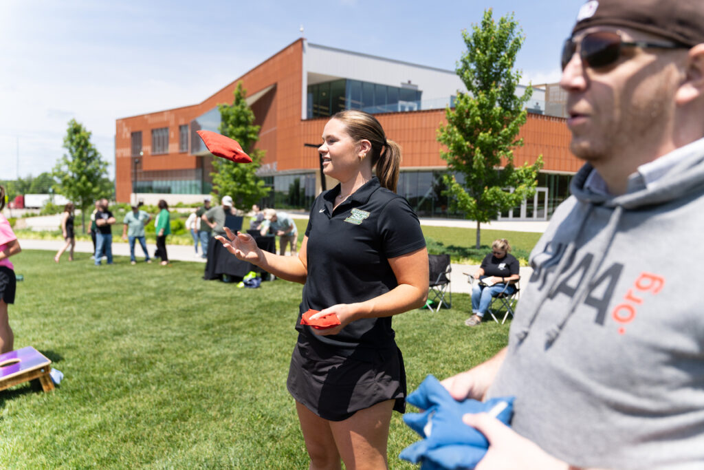 People playing cornhole on a grassy lawn outside a campus building with others in the background.