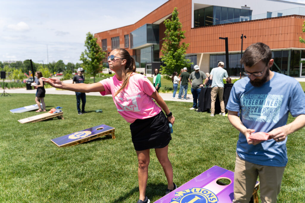 Person playing cornhole on a grassy lawn outside a campus building with others in the background.
