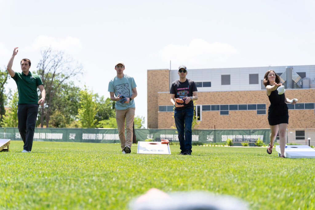 People playing cornhole on a grassy lawn outside a campus building with others in the background.
