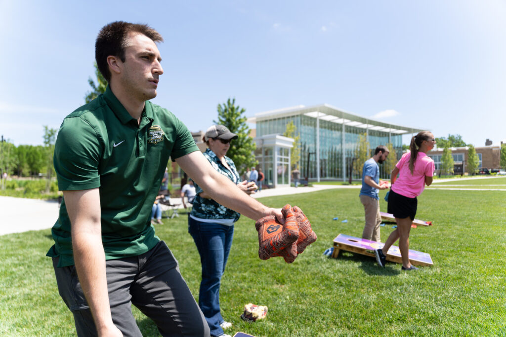 Person playing cornhole on a grassy lawn outside a campus building with others in the background.
