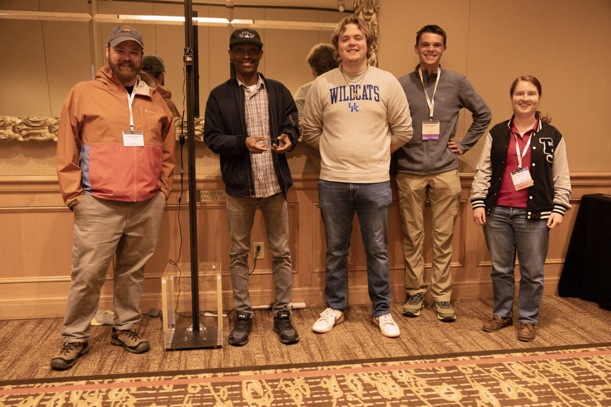 Five people stand side by side indoors at an event, wearing name badges and casual clothing, posing in front of a paneled wall with a display behind them.