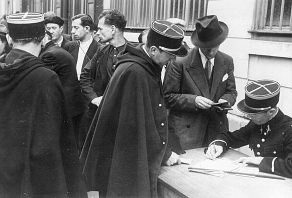 Black-and-white photo of several people in coats and caps gathered around a table, where an official is checking papers and writing, with a building wall and windows in the background.