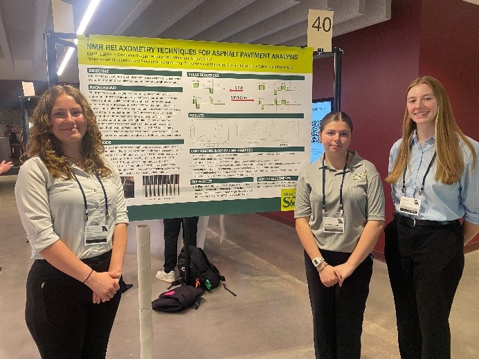Three students wearing conference lanyards stand in front of a research poster.
