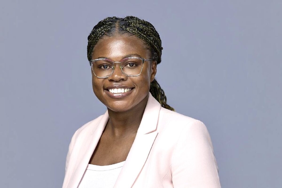 Person wearing glasses and a light pink blazer, smiling in a studio portrait against a gray background.