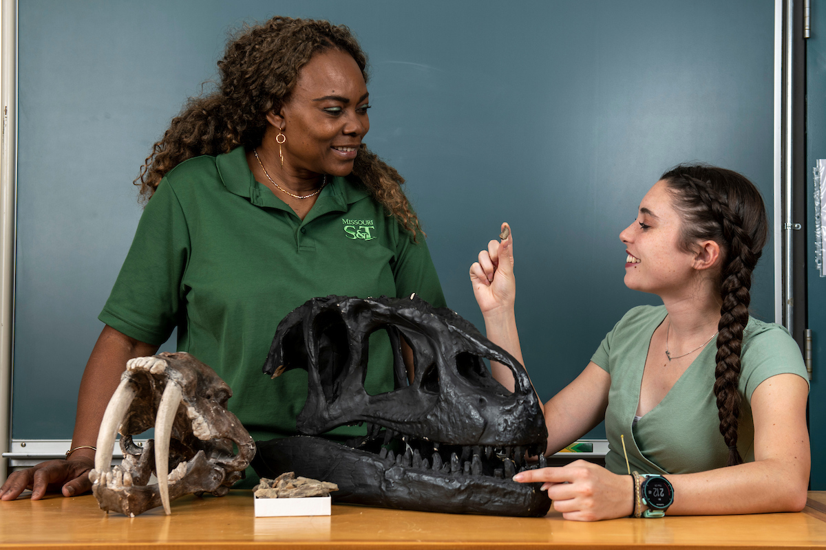 Two individuals examine dinosaur skull replicas on a classroom table, including a large black skull and smaller fossils.