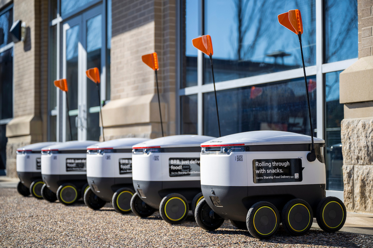 A line of small autonomous delivery robots parked on a sidewalk outside a building.