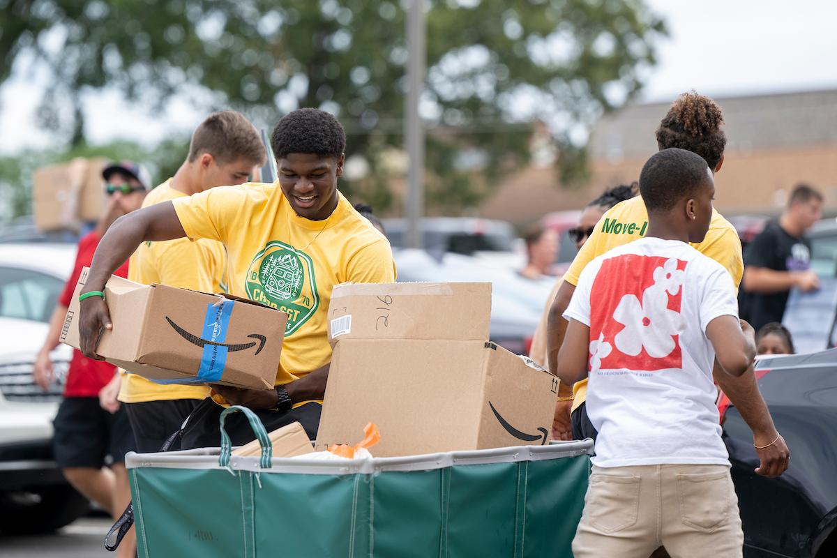 Several individuals load cardboard boxes into a large green cart in a parking lot, some wearing bright yellow shirts, with cars and trees in the background.