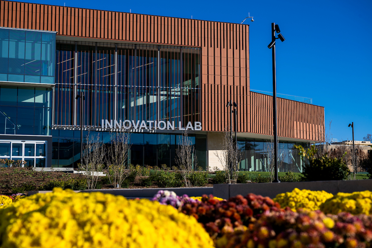 Exterior view of a modern building labeled “Innovation Lab,” featuring large glass windows, vertical metal paneling, and landscaped flower beds in the foreground under a clear blue sky.