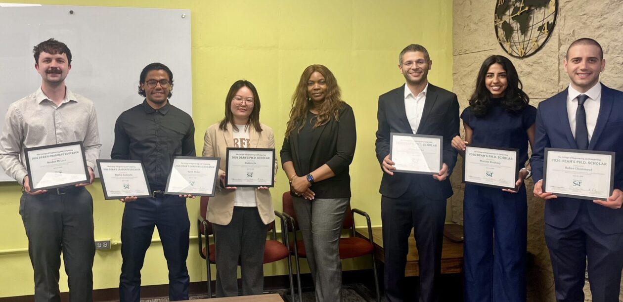 Six people standing indoors against a yellow wall, each holding a framed certificate, with chairs and a table visible in the background.
