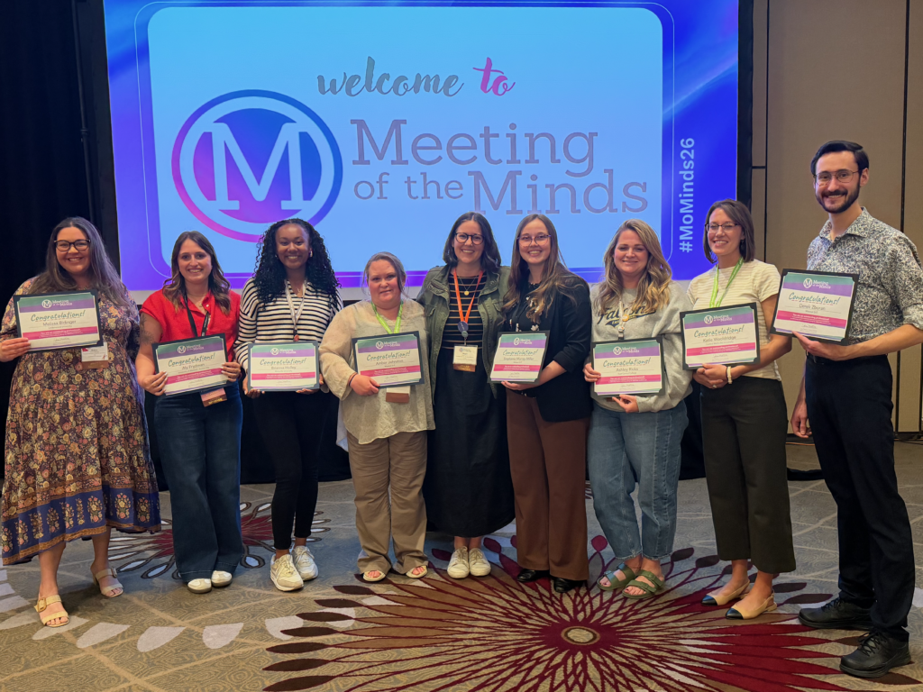 Group of eight people standing at a conference event, each holding a certificate in front of a screen that reads “Welcome to Meeting of the Minds.”