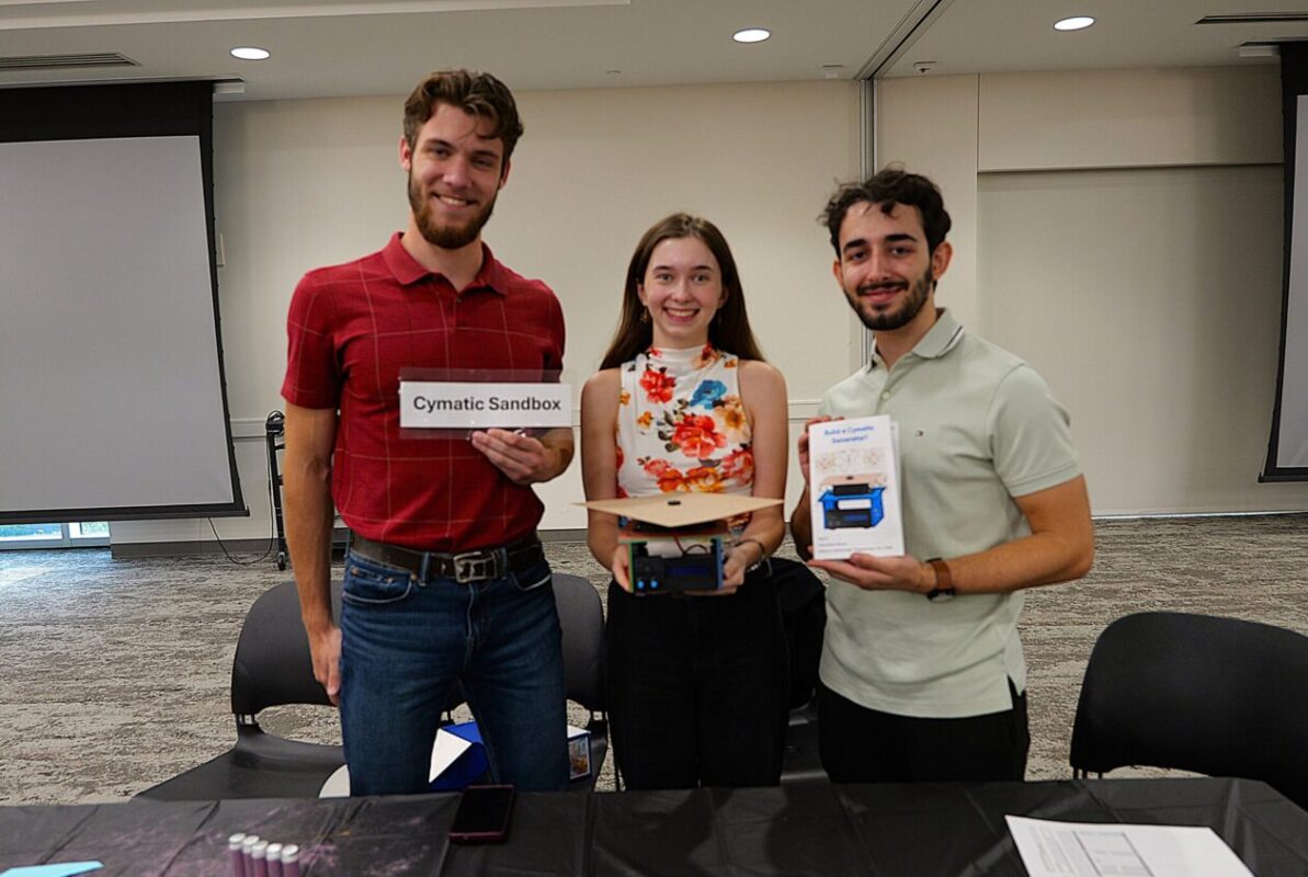 Three people standing behind a table in a conference room, holding product boxes and a sign that reads “Cymatic Sandbox.”
