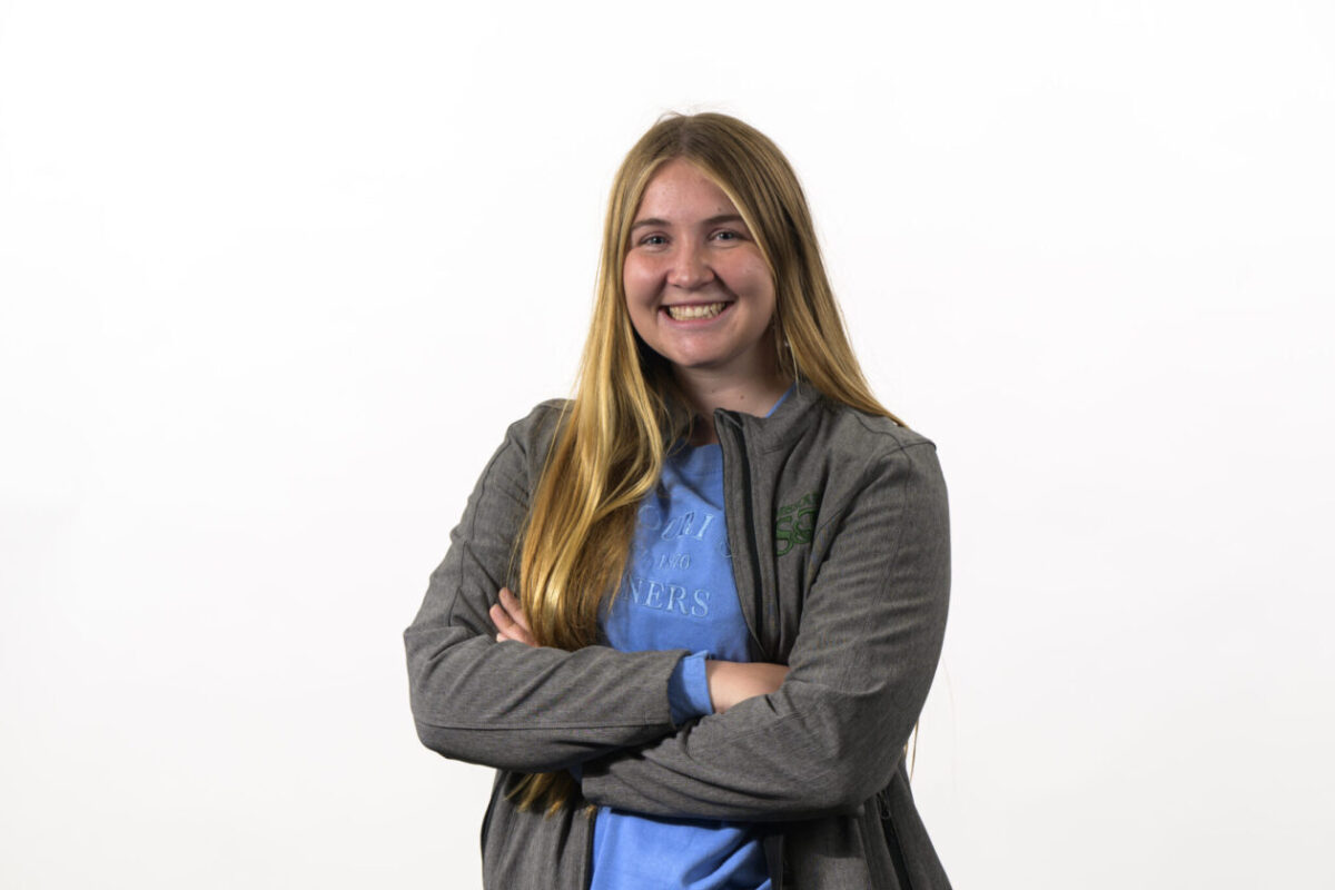Jane Yates stands stands against a plain background, smiling with arms crossed, wearing a blue shirt and gray jacket.