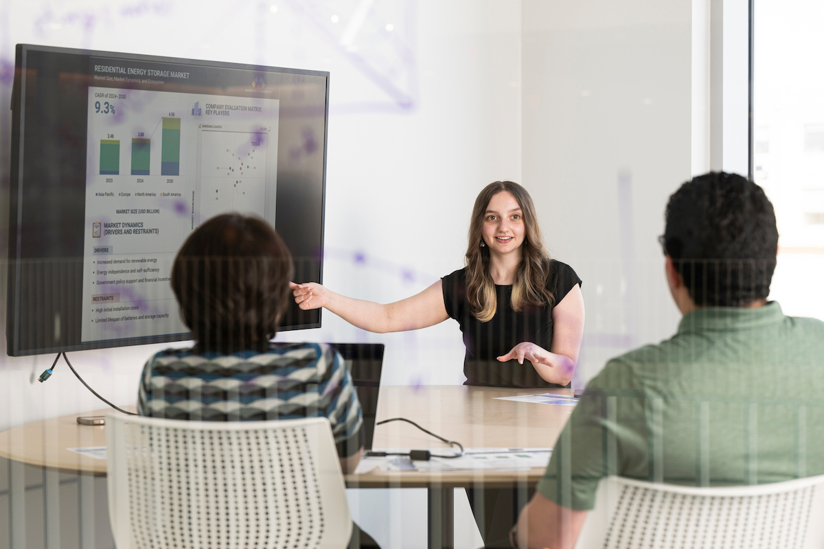 A presenter points to charts and graphs on a display screen while two attendees watch from a conference table.