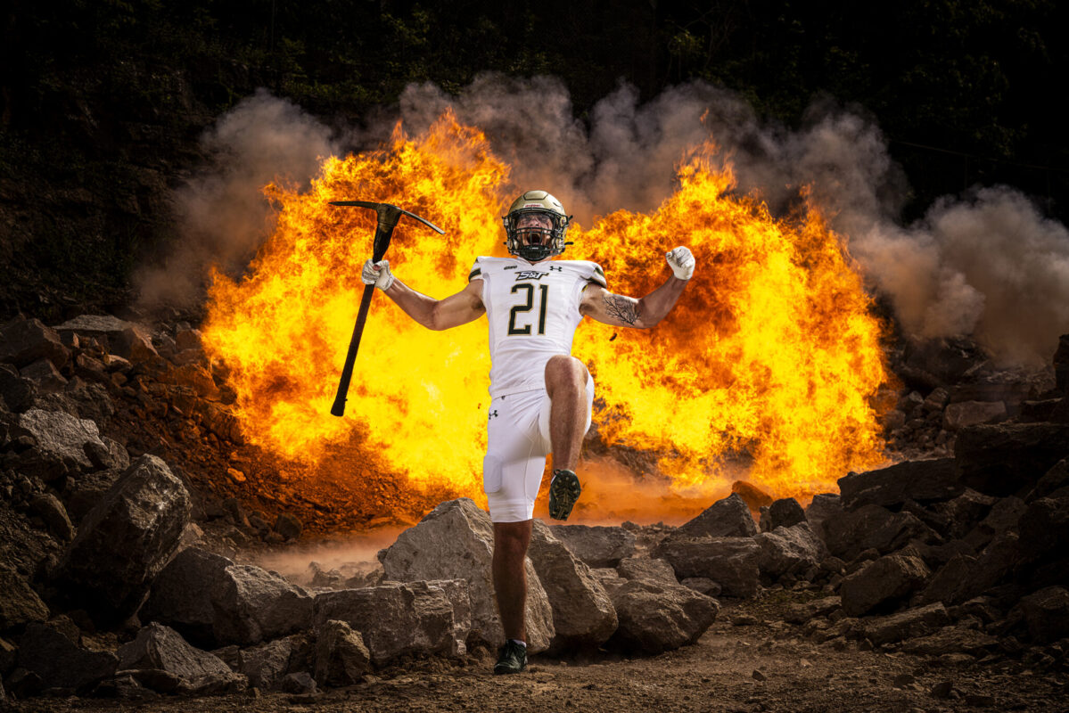 Football player wearing a white uniform with number 21 runs through rocky terrain holding a pickaxe, with a large fireball blazing in the background.
