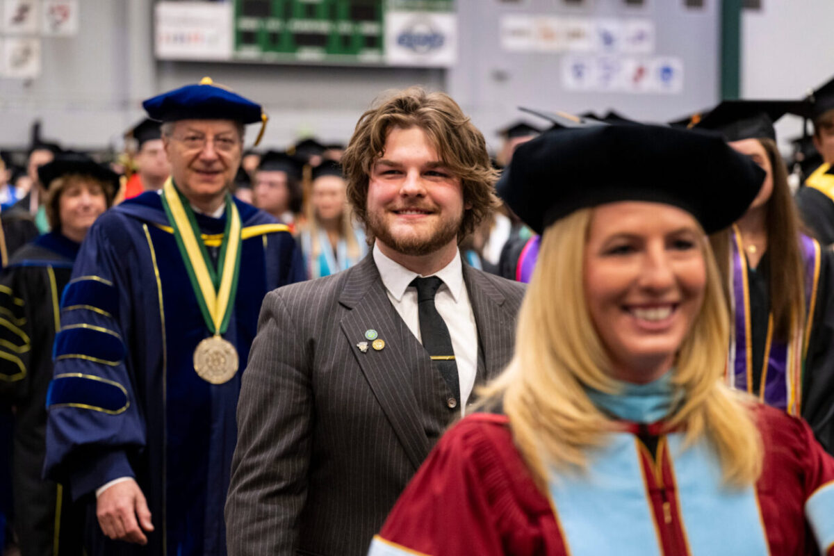 Participants in academic regalia and formal attire process through an indoor commencement ceremony, with university leaders and graduates visible in the background.