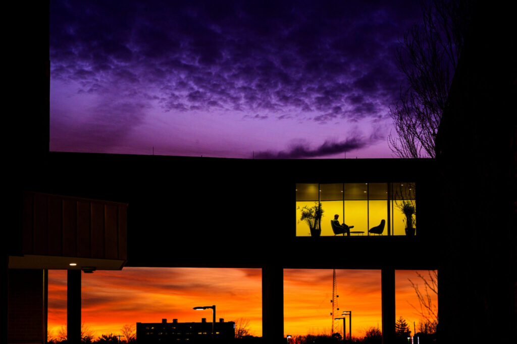 A building is silhouetted against a colorful sunset and twilight sky, with orange and purple hues filling the horizon. Warm light from an upper-floor window reveals indoor seating and plants.