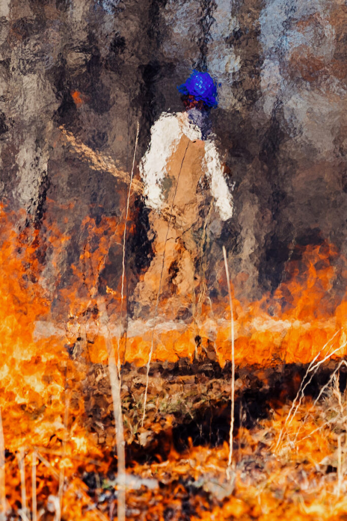 A prescribed burn moves through a grassy woodland area as a firefighter in protective gear walks behind the flames.