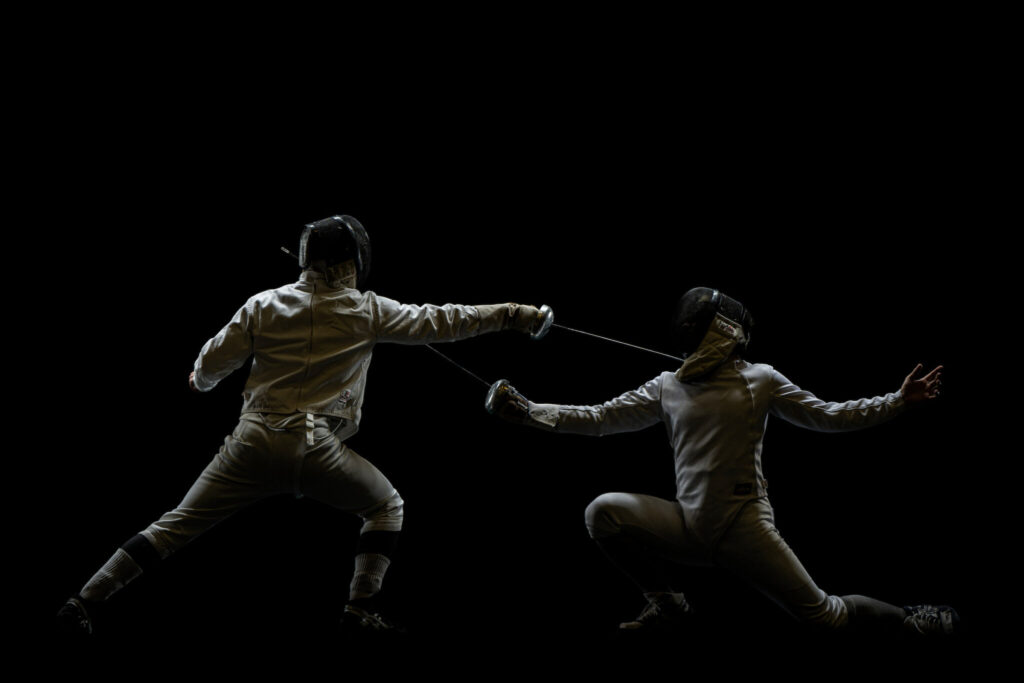 Two fencers in protective gear compete with épées on a dark stage, lunging toward each other during a bout.