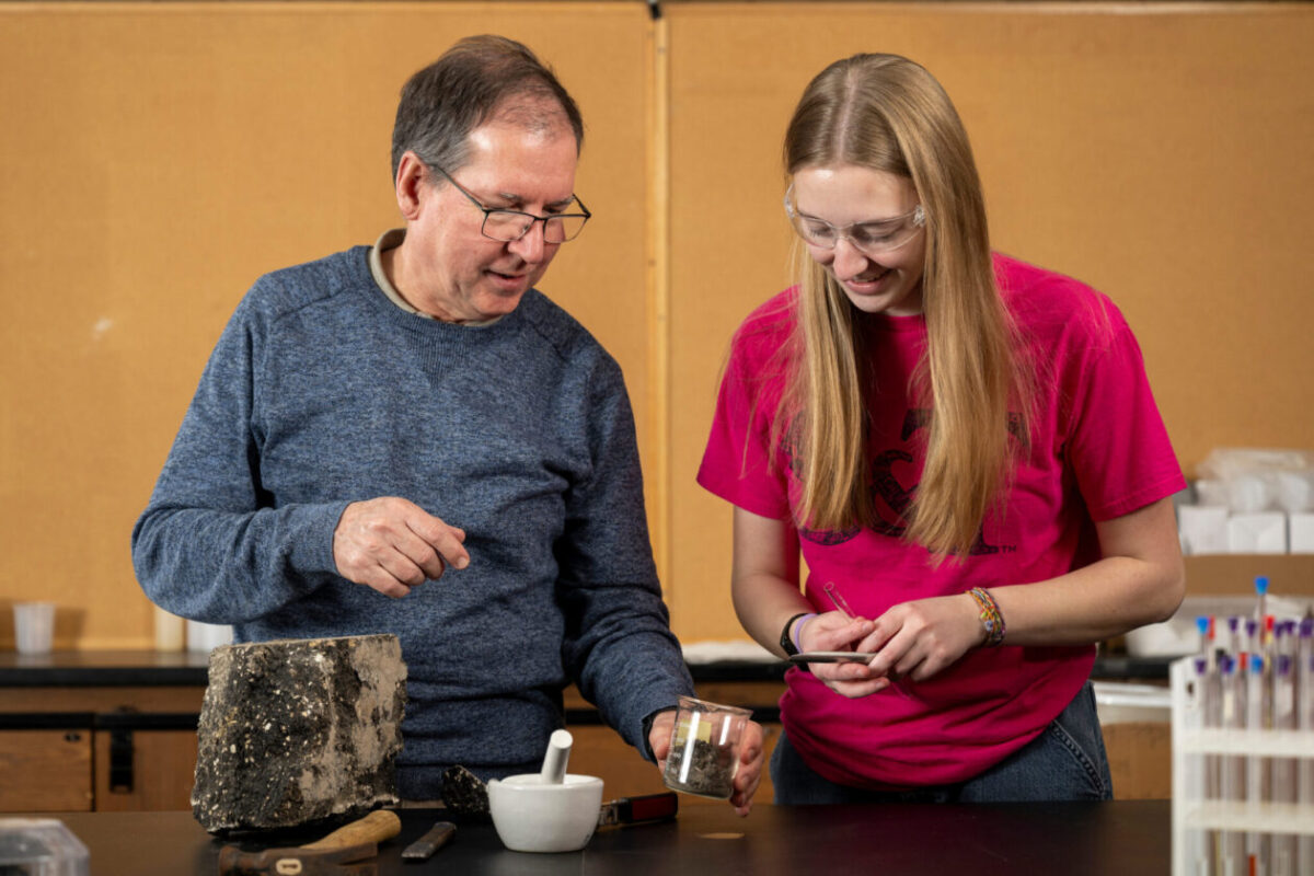 A faculty member and student work together at a laboratory bench, examining a sample container alongside testing equipment and rock specimens.