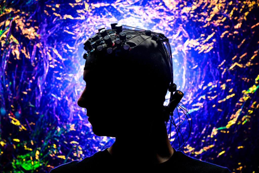 Silhouette of a person wearing a brain-monitoring cap with sensors and wires, set against a vibrant purple, blue and gold illuminated background.