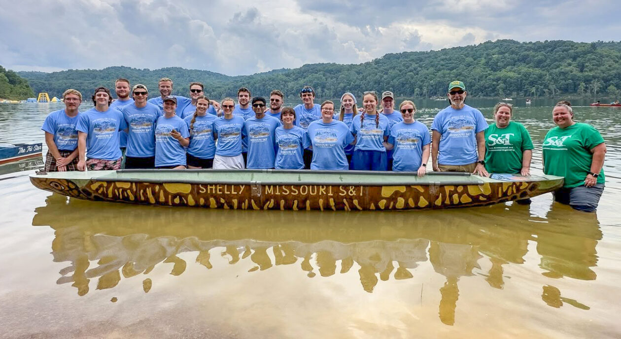A group of students and staff wearing matching blue and green Missouri S&T T-shirts stand in shallow water beside a long canoe on a lake, with forested hills and cloudy skies in the background.